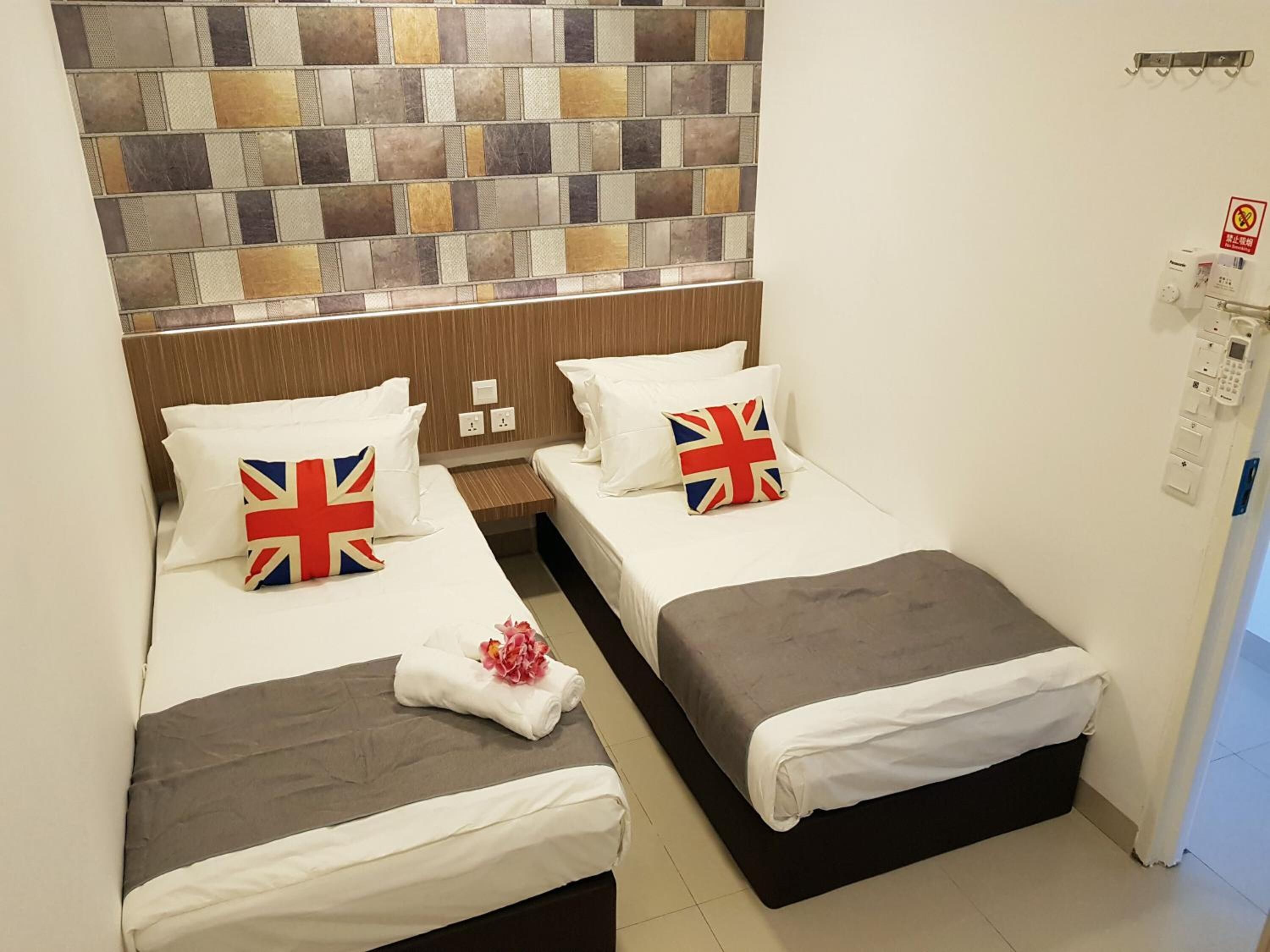 A hotel room with two beds, each with a pillow featuring the Union Jack flag. There is a small table between the beds, and the walls are adorned with a mosaic tile design.