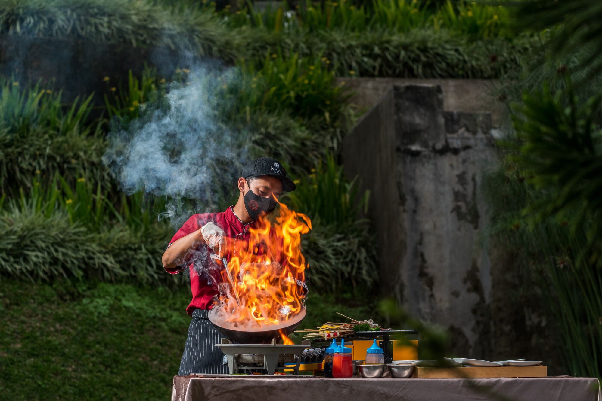 Un chef cocina con llamas, rodeado de una mesa con utensilios de cocina y una parrilla.