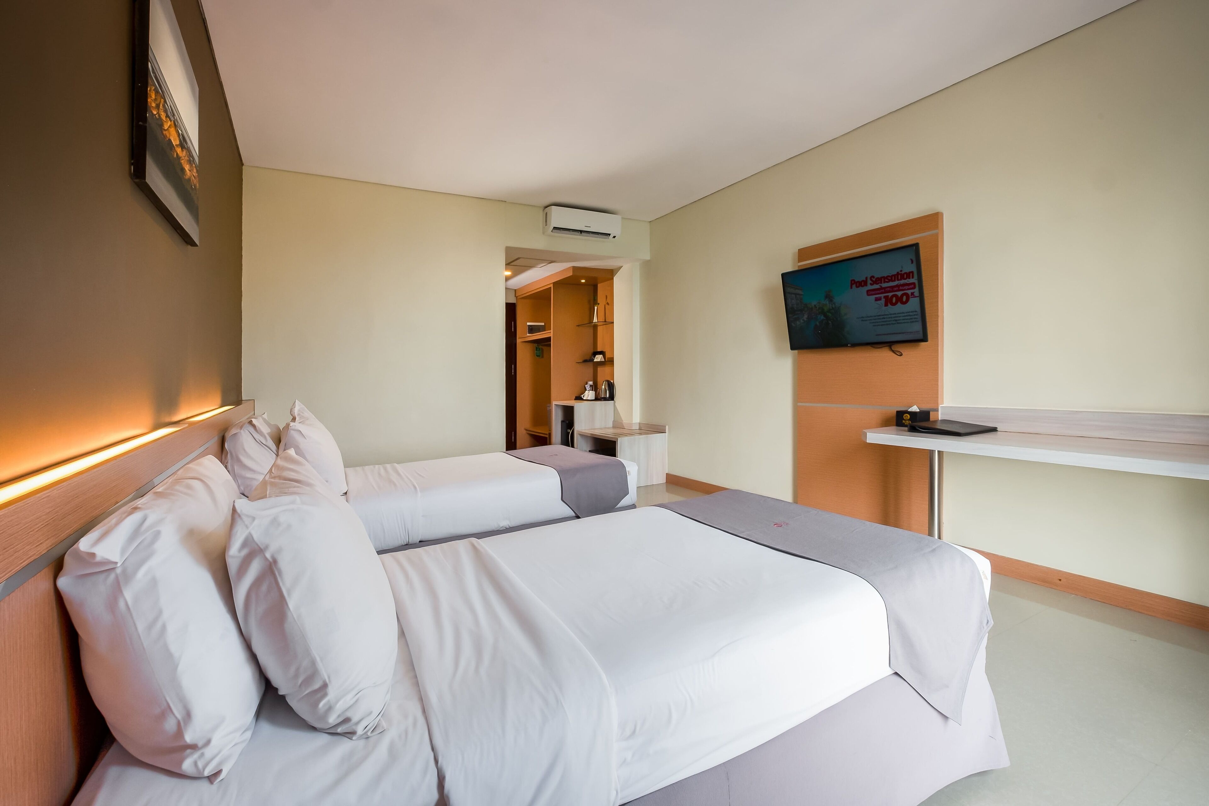 A hotel room with two beds, a TV mounted on the wall, a desk, and a shelf with a book.
