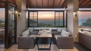Un espace extérieur moderne, aménagé avec des meubles en osier, une table en bois et de grandes fenêtres offrant une vue sur les montagnes.