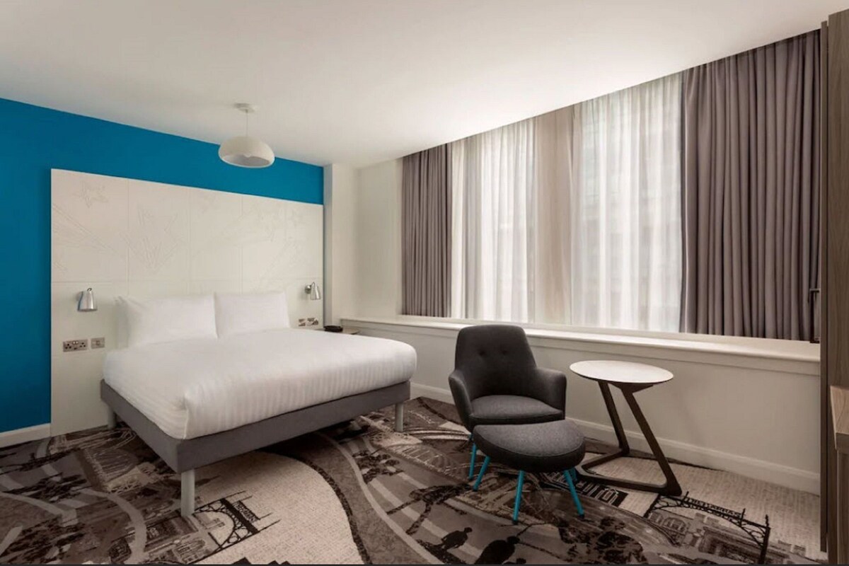 A modern hotel room with a bed, a chair, a small table, and a large window with curtains.