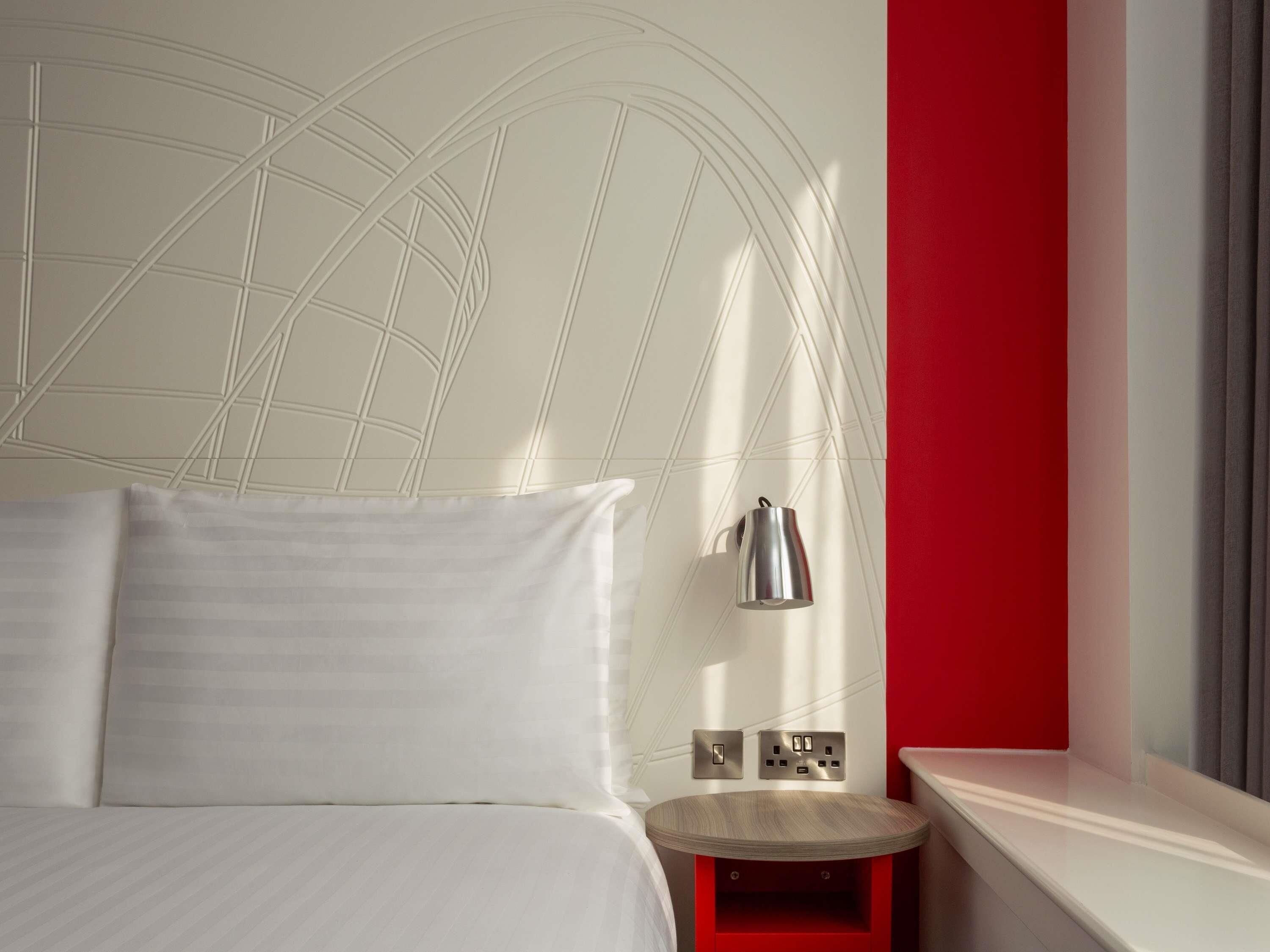 A neatly made bed with white linens, a wall-mounted lamp, and a small wooden side table with a red accent.