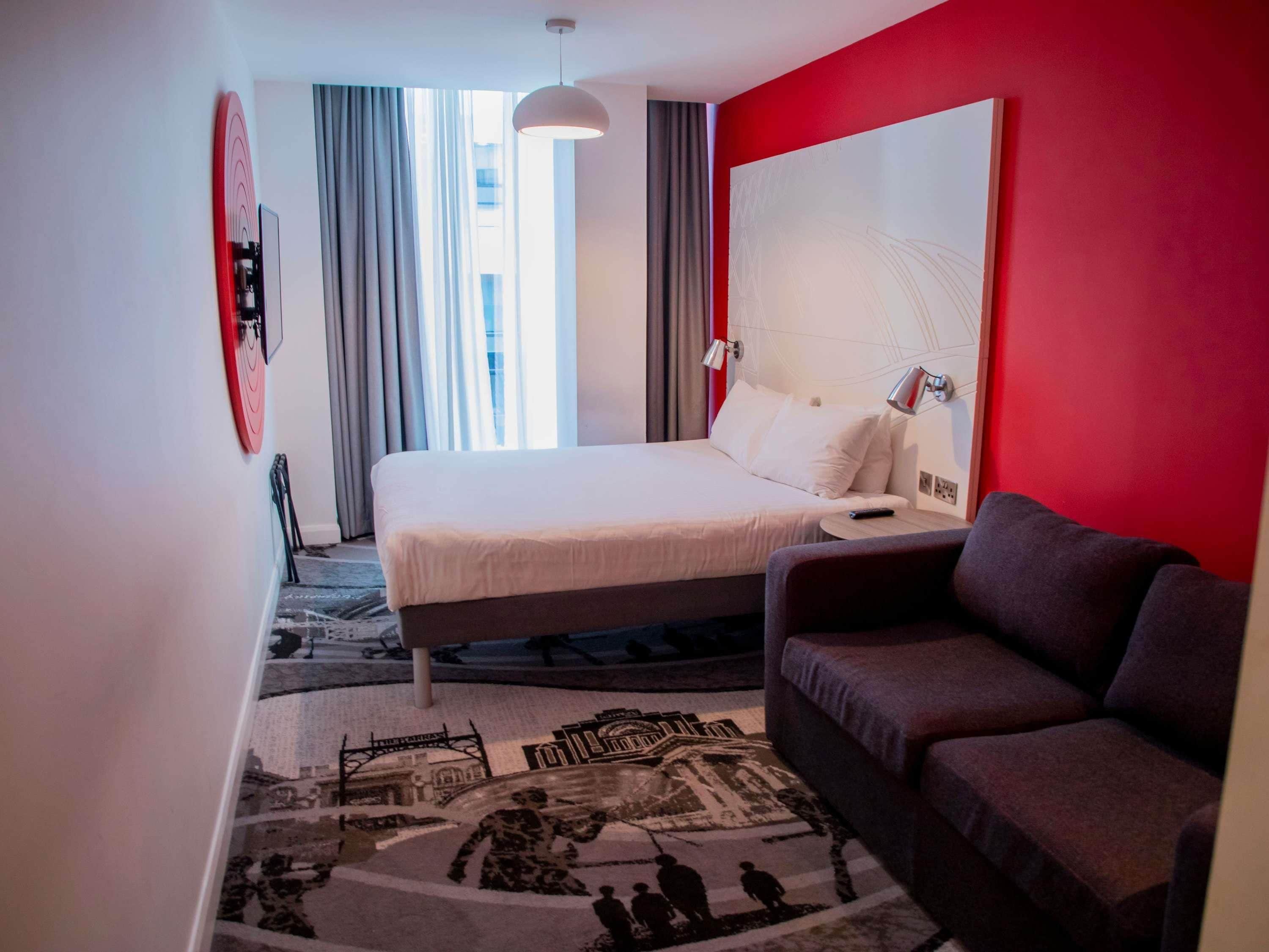 A hotel room with a bed, a sofa, a large mirror, a nightstand, and a window with curtains.