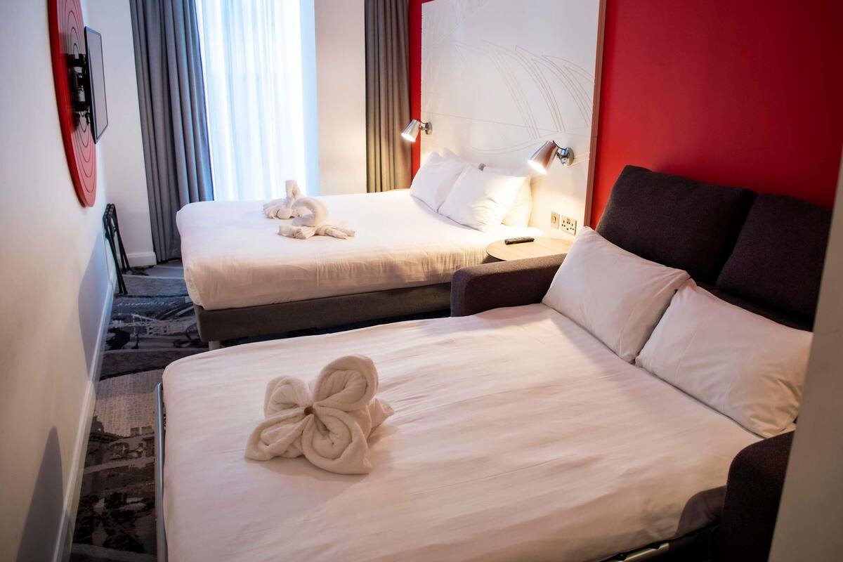 A hotel room with two beds, a red accent wall, a large abstract artwork, and a view of the outdoors.
