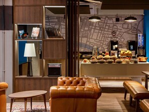A modern hotel lobby with a leather sofa, a small table, a lamp, a map wall, and a buffet area. - Ibis Styles Glasgow Central (Glasgow)