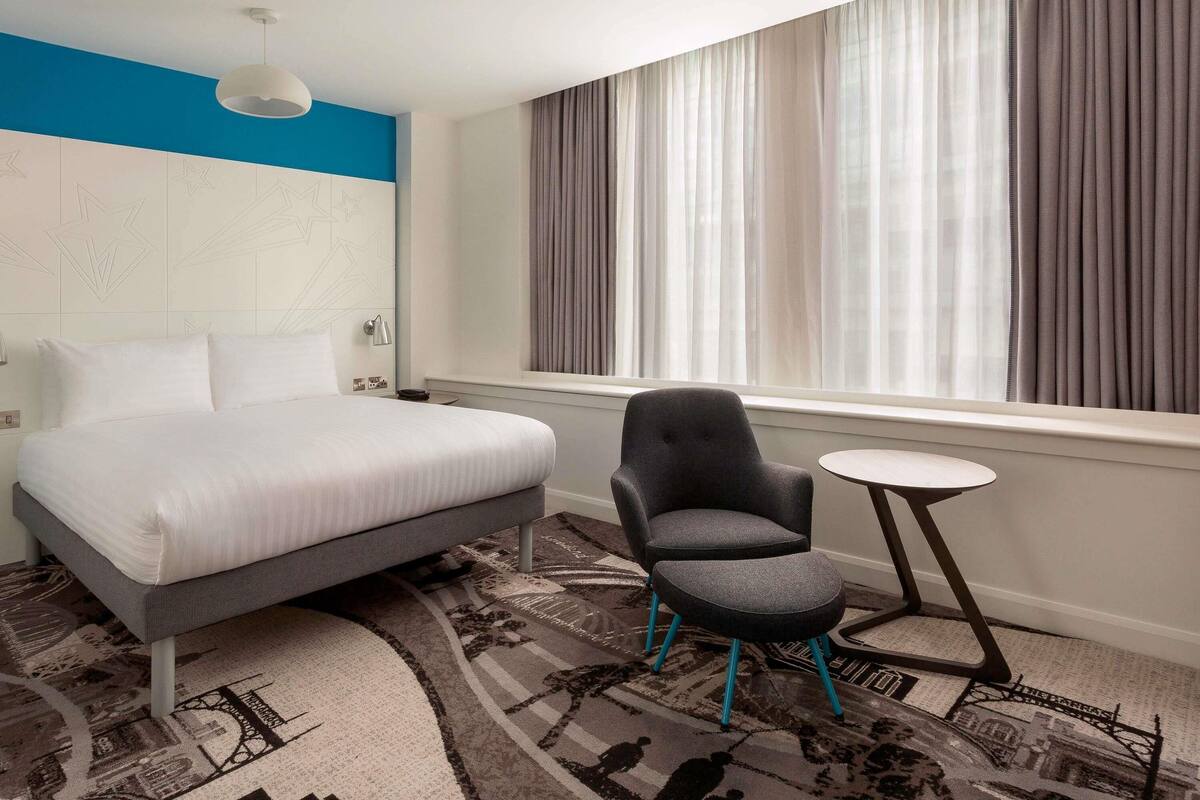 A hotel room with a bed, a chair, a small table, a window with curtains, and a patterned carpet.
