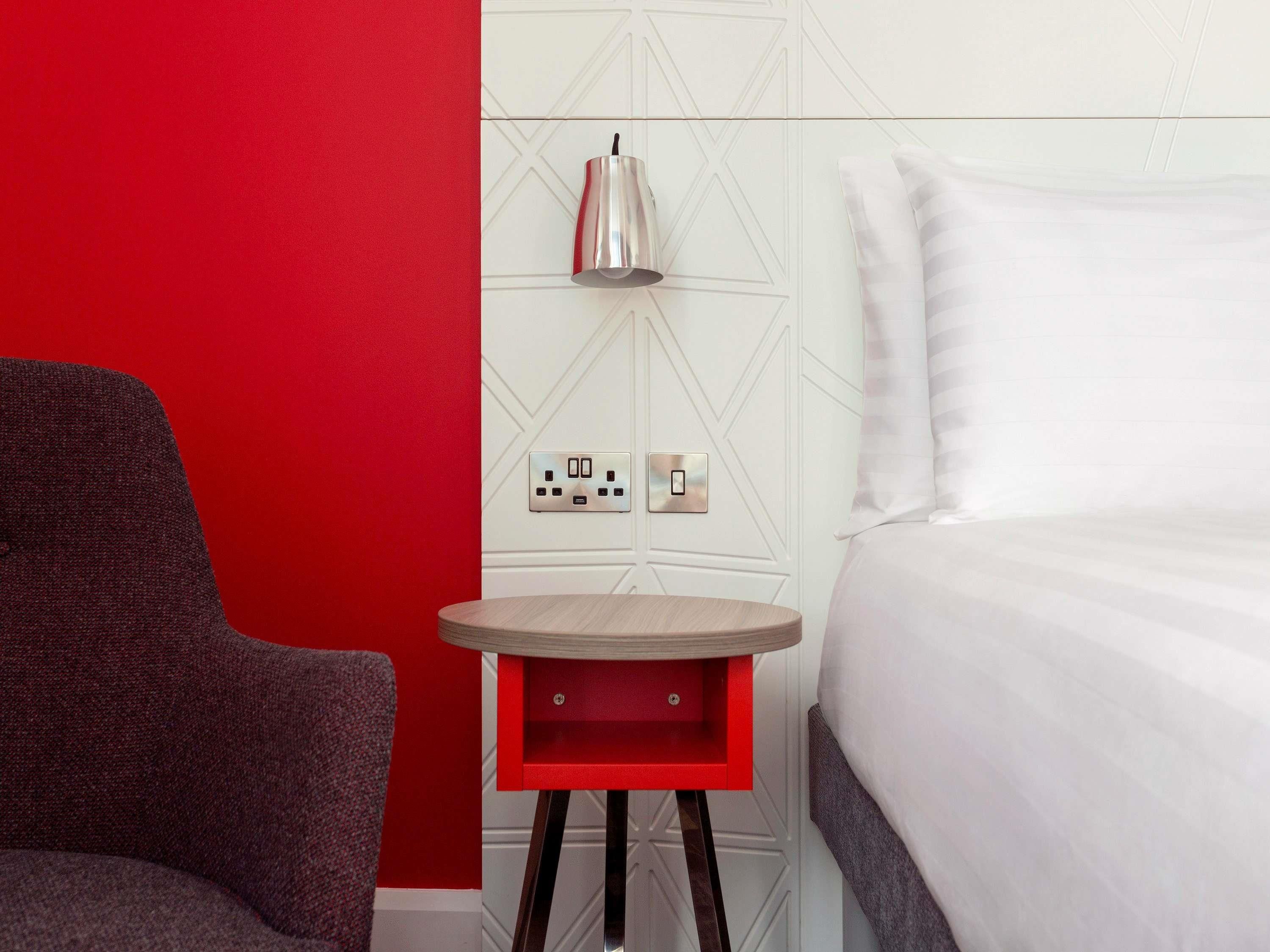 A hotel room with a red wall, a bed with white bedding, a small table with a lamp, and a chair.