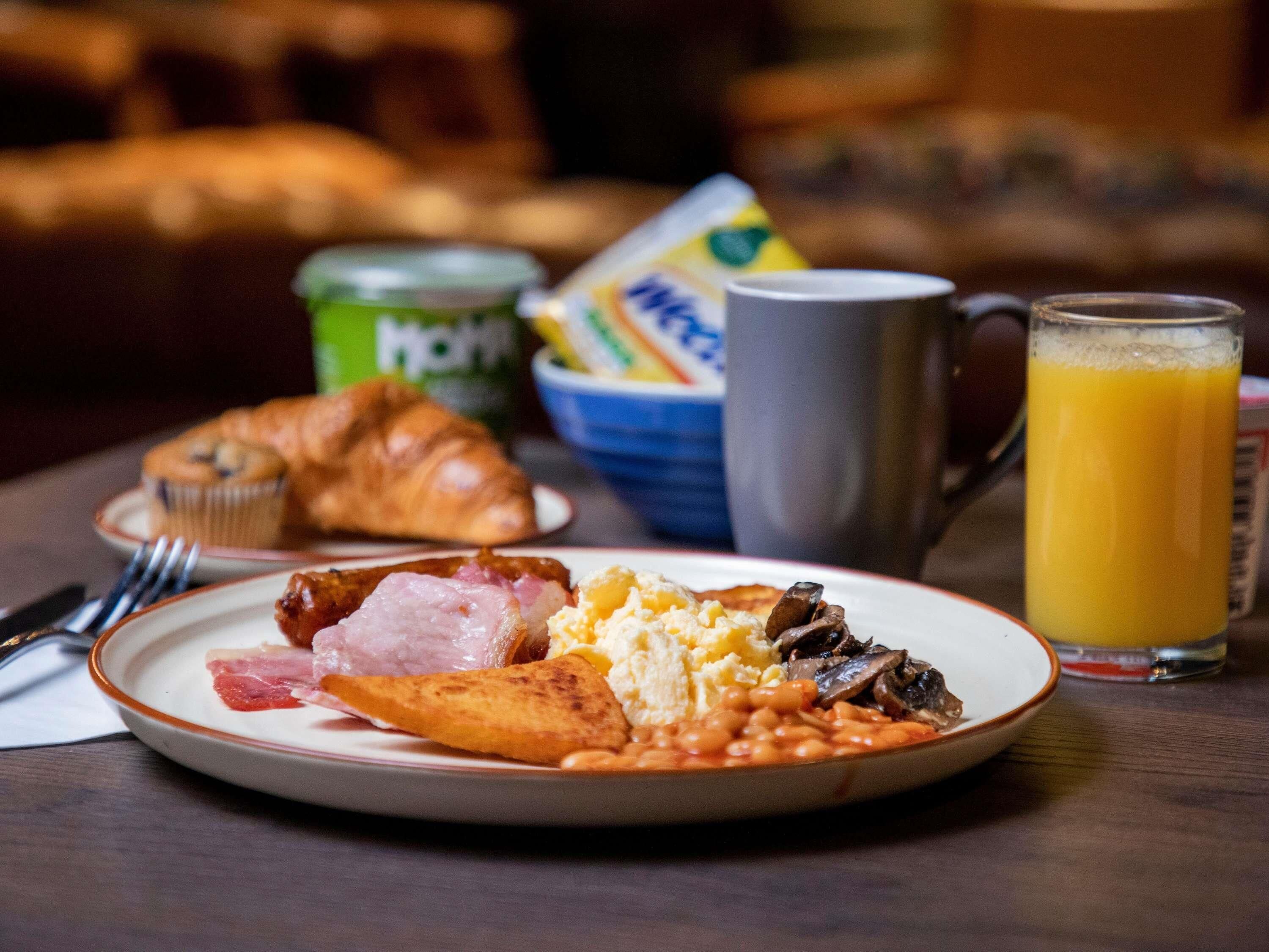A breakfast plate with eggs, beans, ham, and a croissant, a mug, and a glass of orange juice on a table.