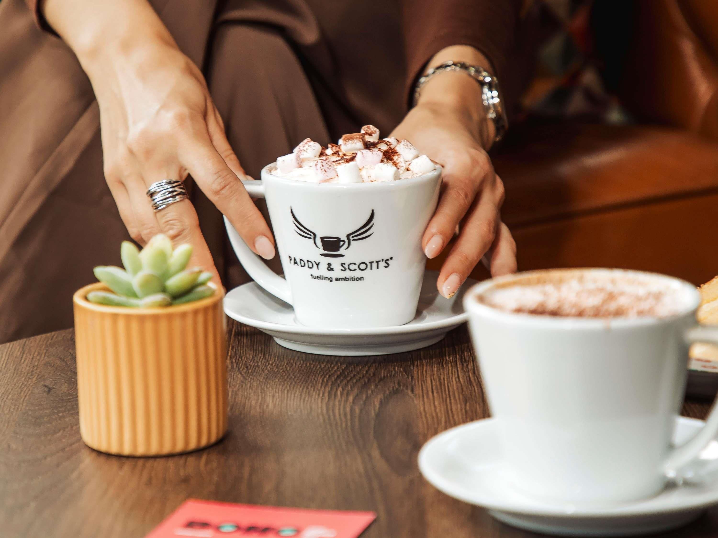A person holding a cup with 'FADDY & SCOTT'S' logo, a small potted plant, and a cup of hot beverage on a wooden table.
