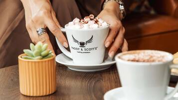 A person holding a cup with 'FADDY & SCOTT'S' logo, a small potted plant, and a cup of hot beverage on a wooden table.