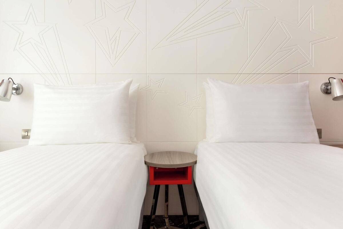A hotel room with two beds, a small red table, and wall-mounted lamps.