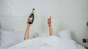 A person holding a bottle and a glass in a bed with white bedding and a geometric wall design. - Ibis Styles Glasgow Central (Glasgow)