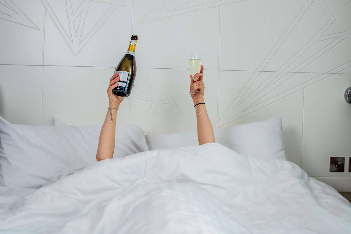 A person holding a bottle and a glass in a bed with white bedding and a geometric wall design.