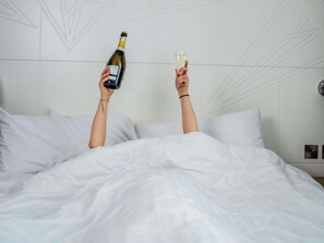 Uma pessoa segurando uma garrafa e um copo em uma cama com roupa de cama branca e um design geométrico na parede.