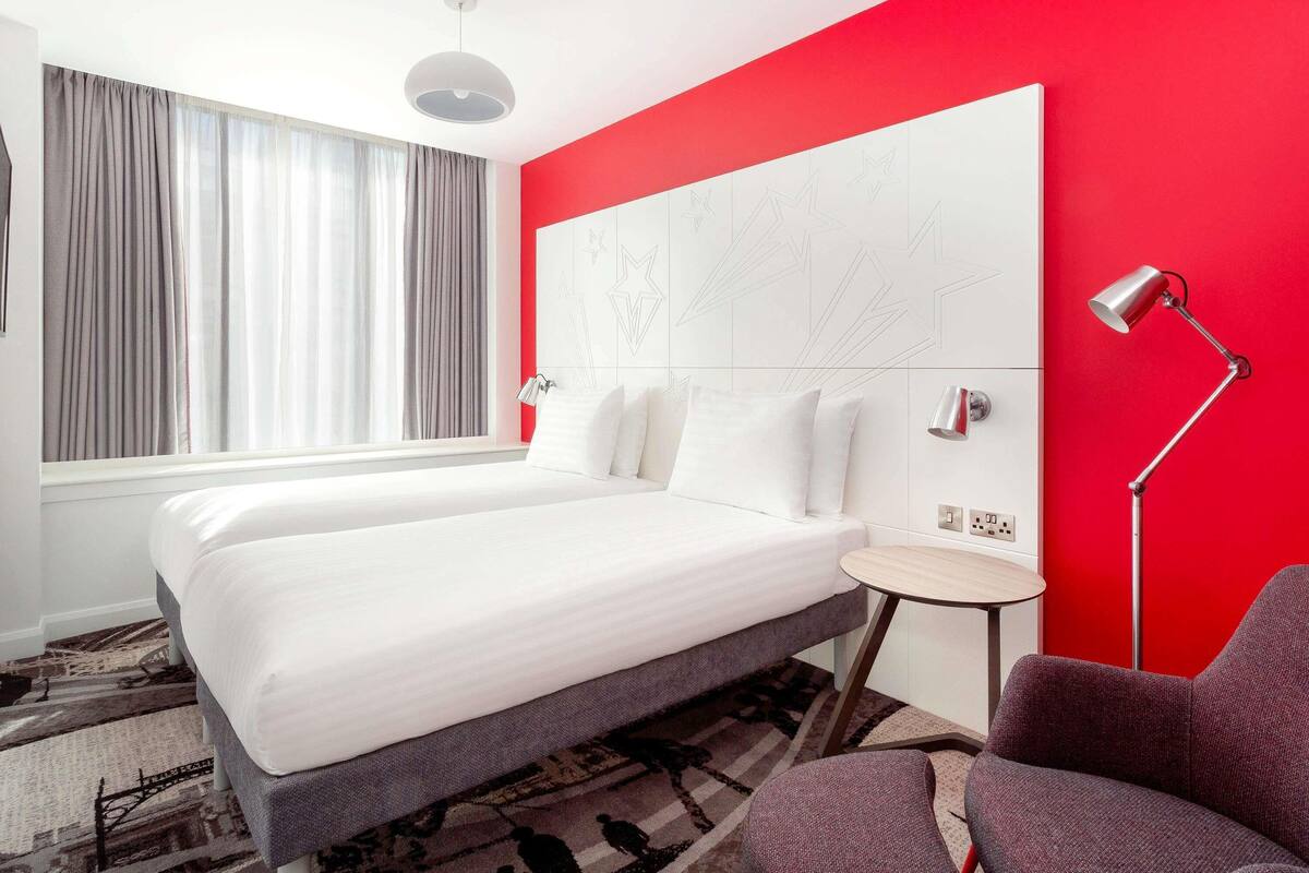 A hotel room with a large bed, a small table, a chair, a lamp, and a window with curtains.