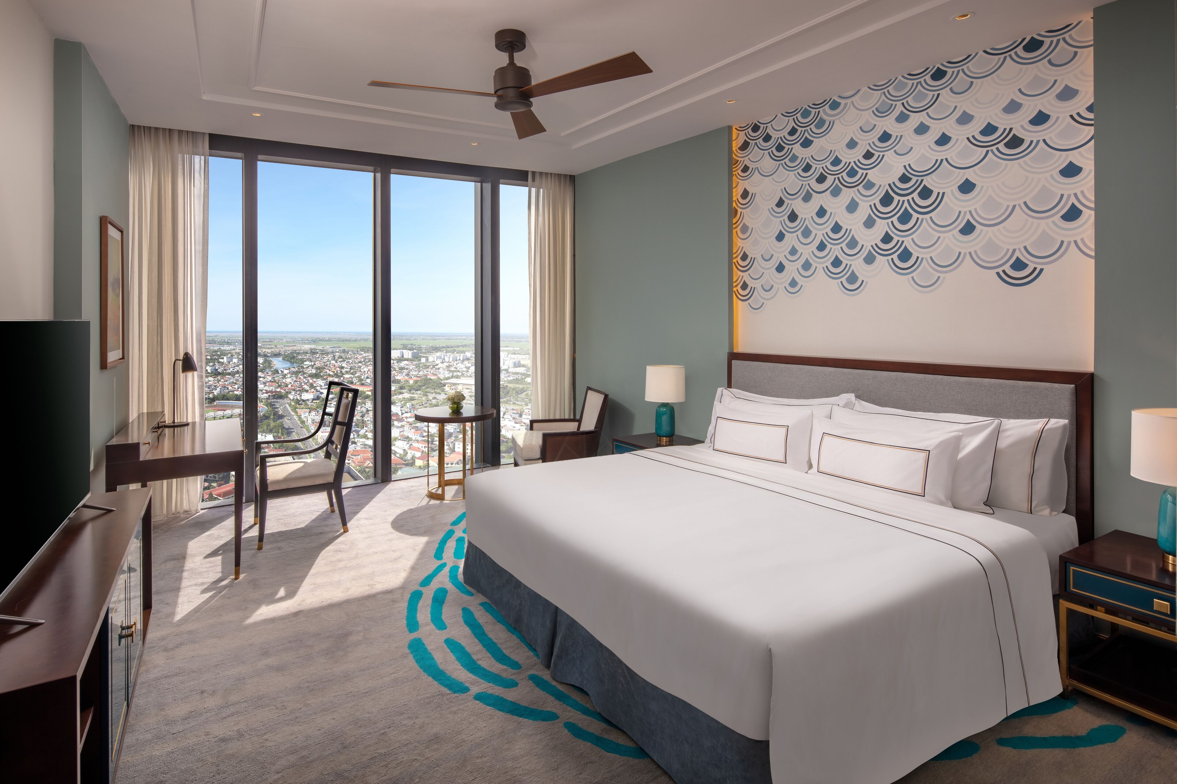 A hotel room with a large bed, a desk, a chair, a ceiling fan, and a view of the city through large windows.