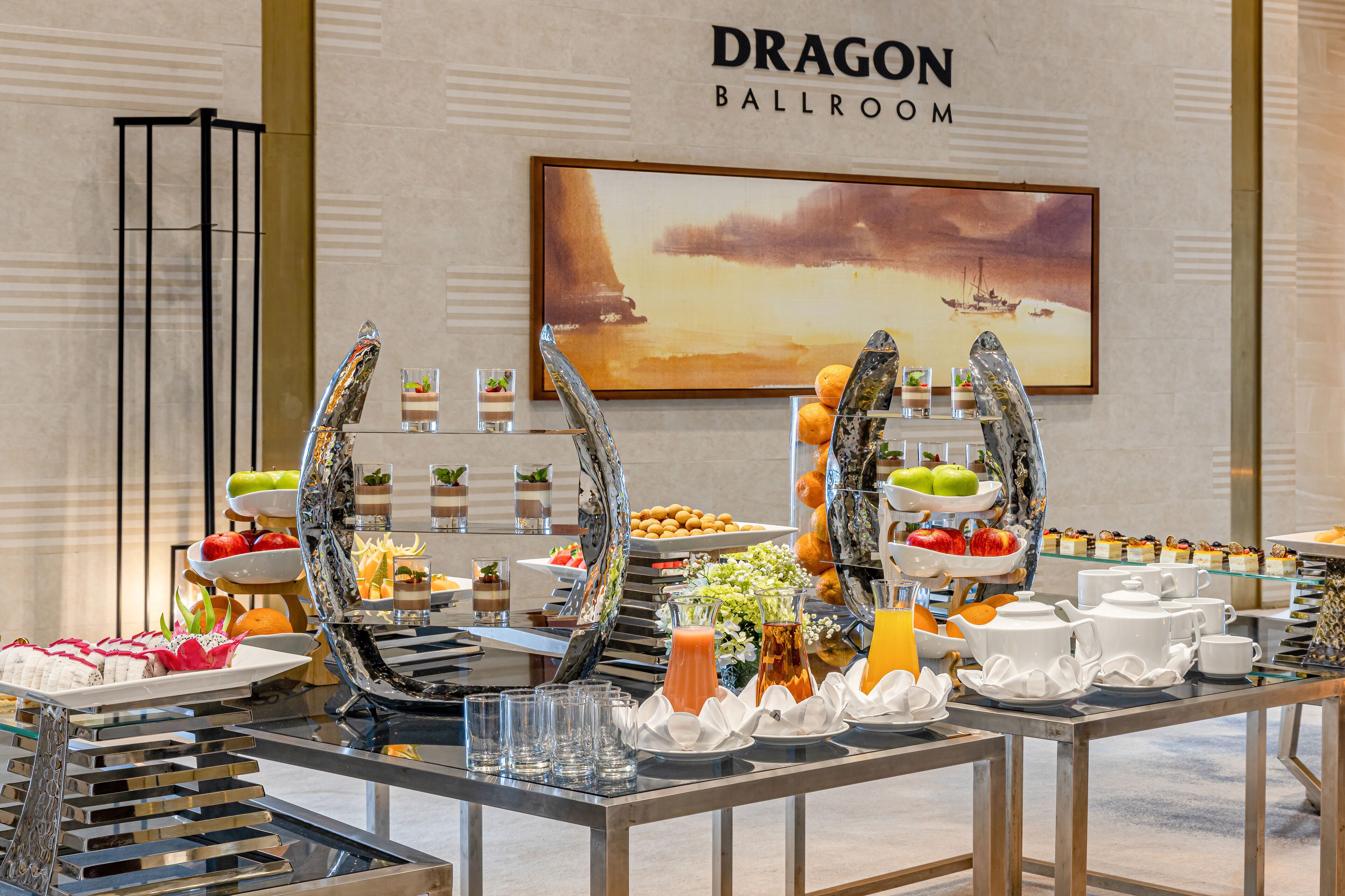 A buffet setup with various food items and beverages, a large painting of a sunset, and a sign reading "DRAGON BALLROOM".