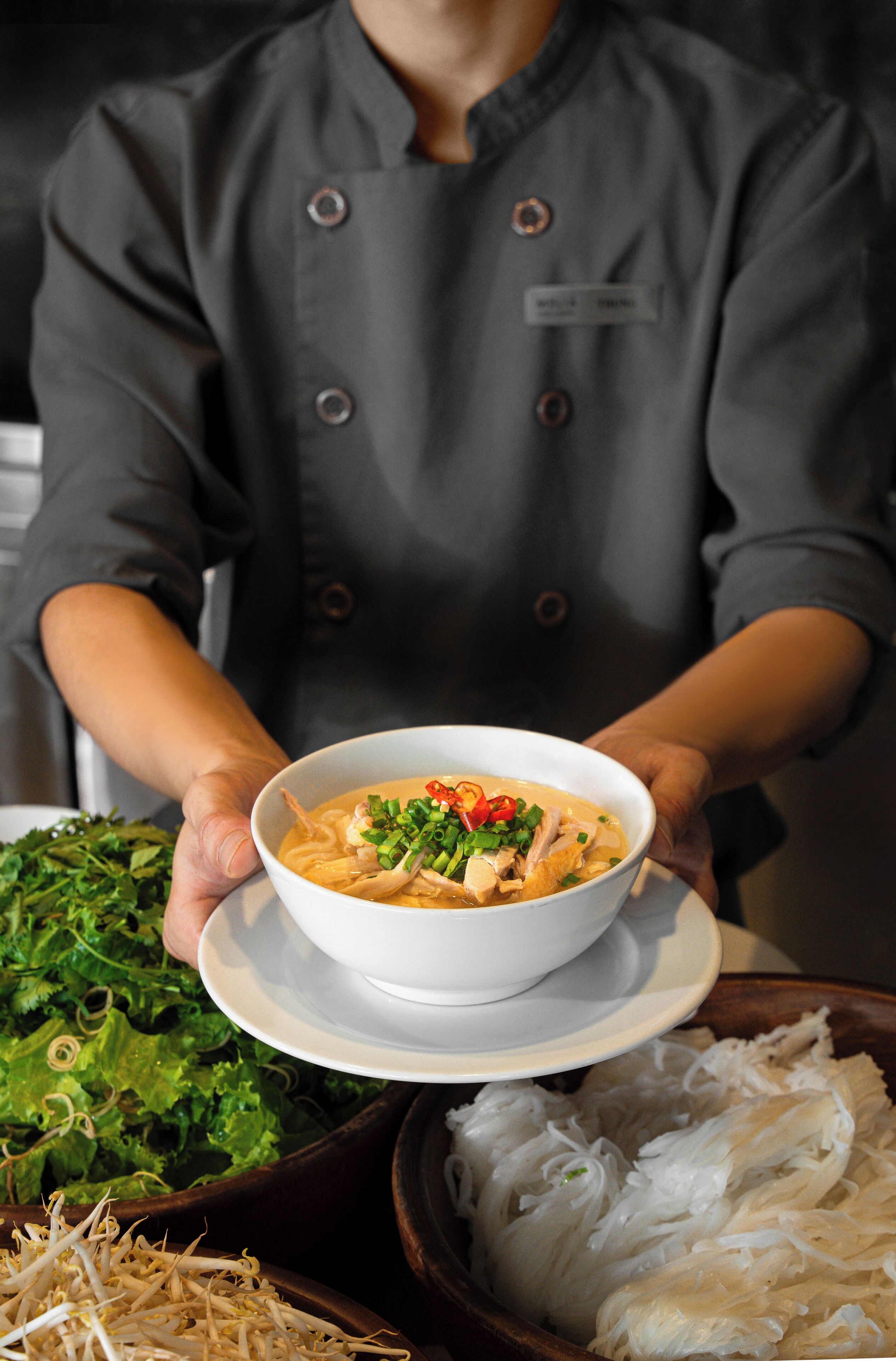 Un chef sosteniendo un bol de sopa con un plato de verduras y fideos.