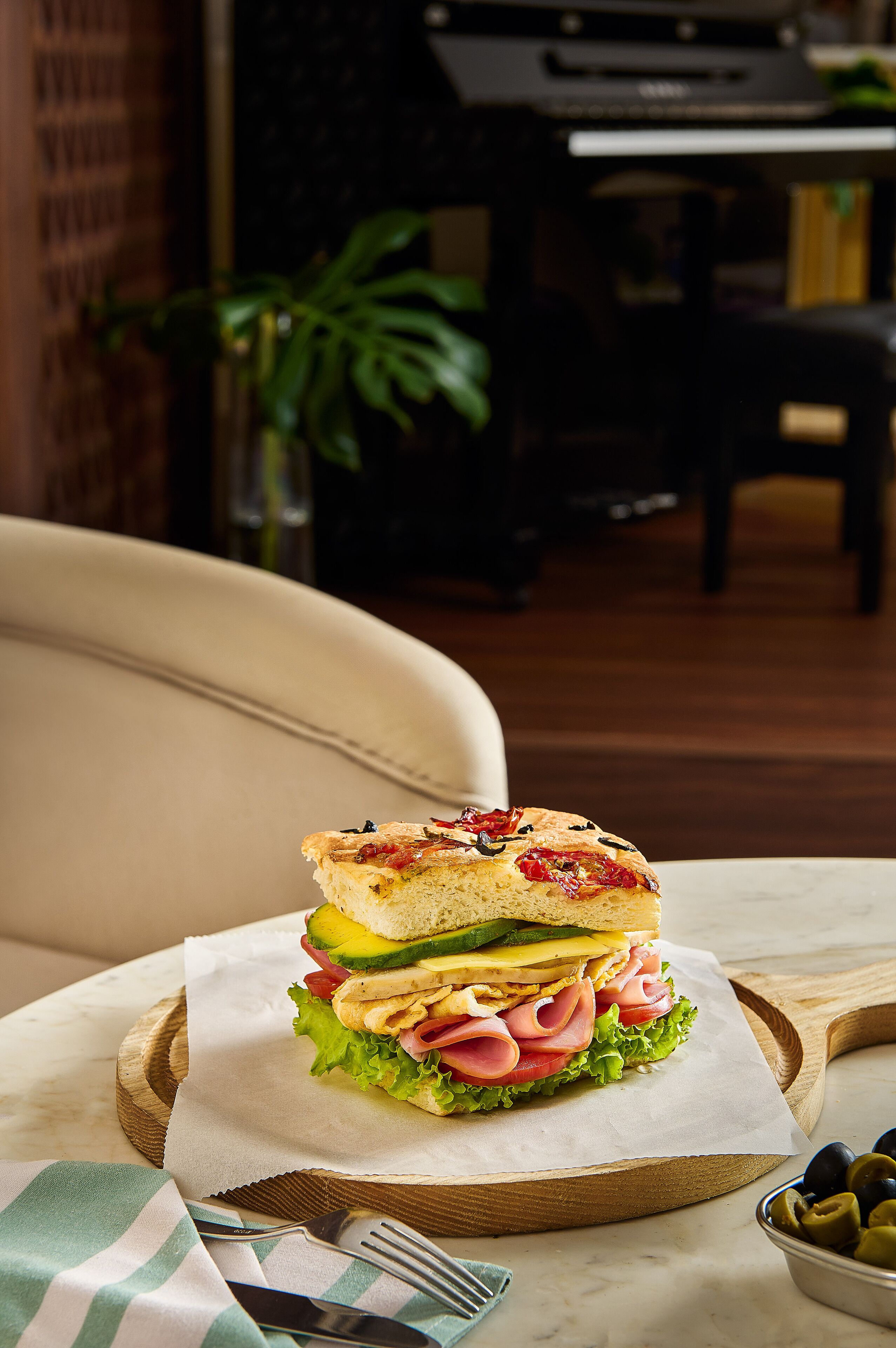 A sandwich on a wooden board with a napkin, a fork, and a bowl of olives on a table.