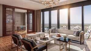 Un vaste salon doté d’une grande fenêtre offrant une vue sur la ville, d’un lustre moderne et d’un coin salon confortable avec un canapé et des fauteuils.