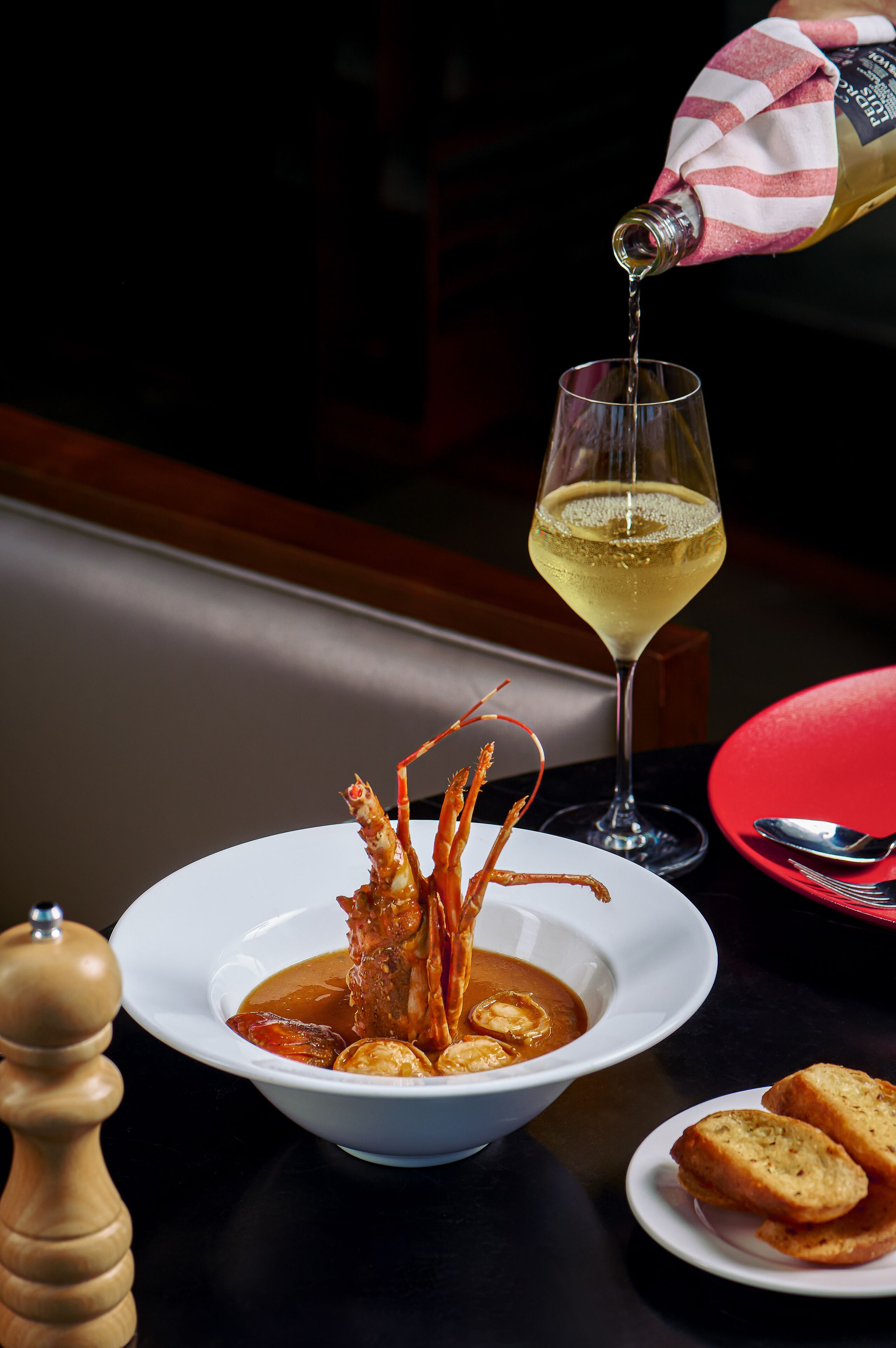 A bowl of seafood soup with a lobster tail, a glass of white wine, and bread on a table.