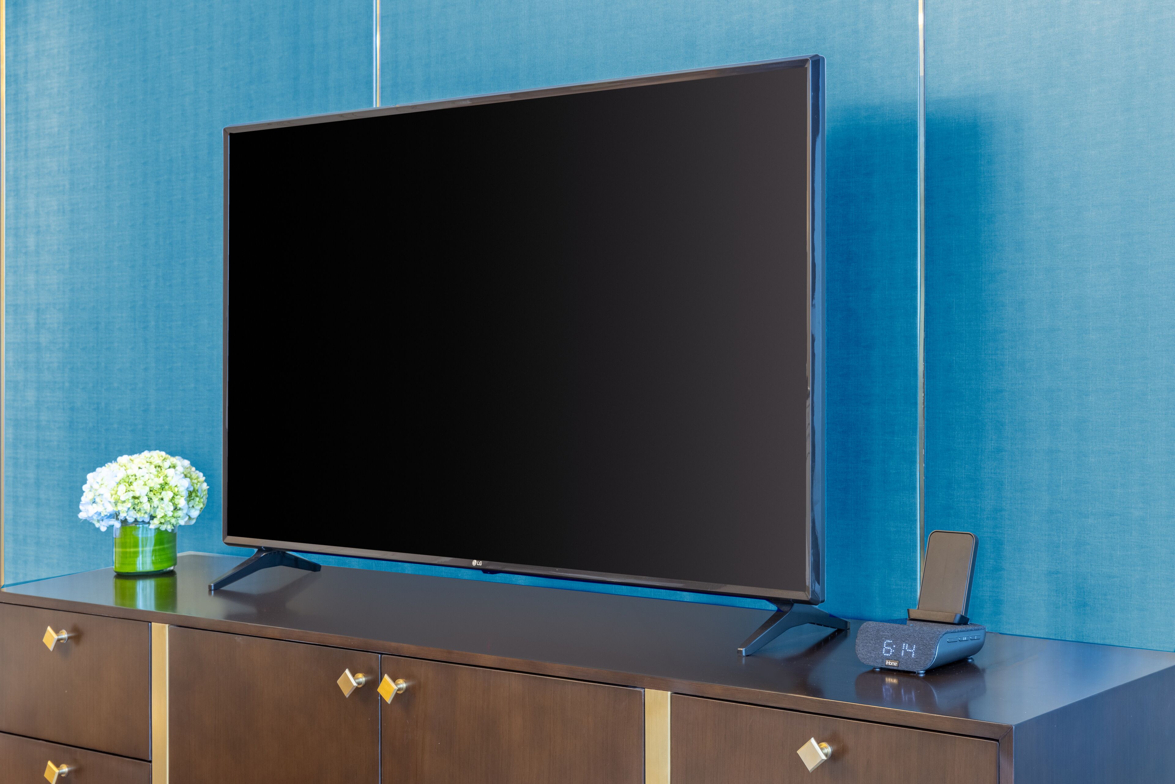 Un televisor de pantalla plana moderno sobre un aparador de madera con un jarrón de flores y un reloj digital.