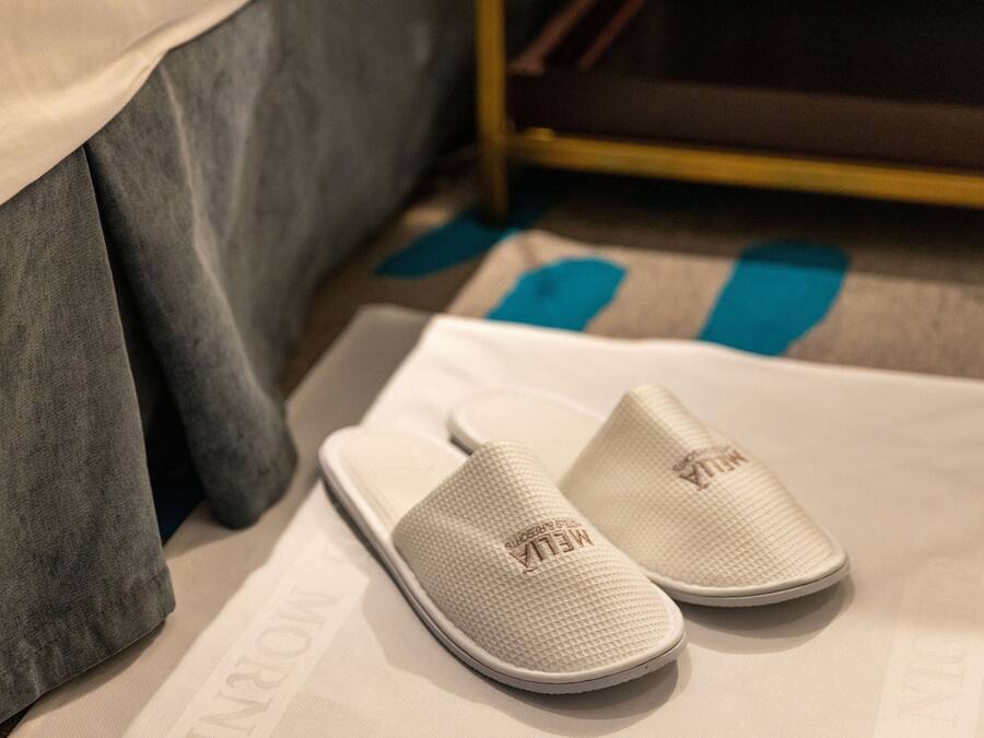 Un par de pantuflas blancas con un diseño texturizado y la palabra “MELT” en la suela, sobre una cama con una manta gris y un objeto azul en el suelo.