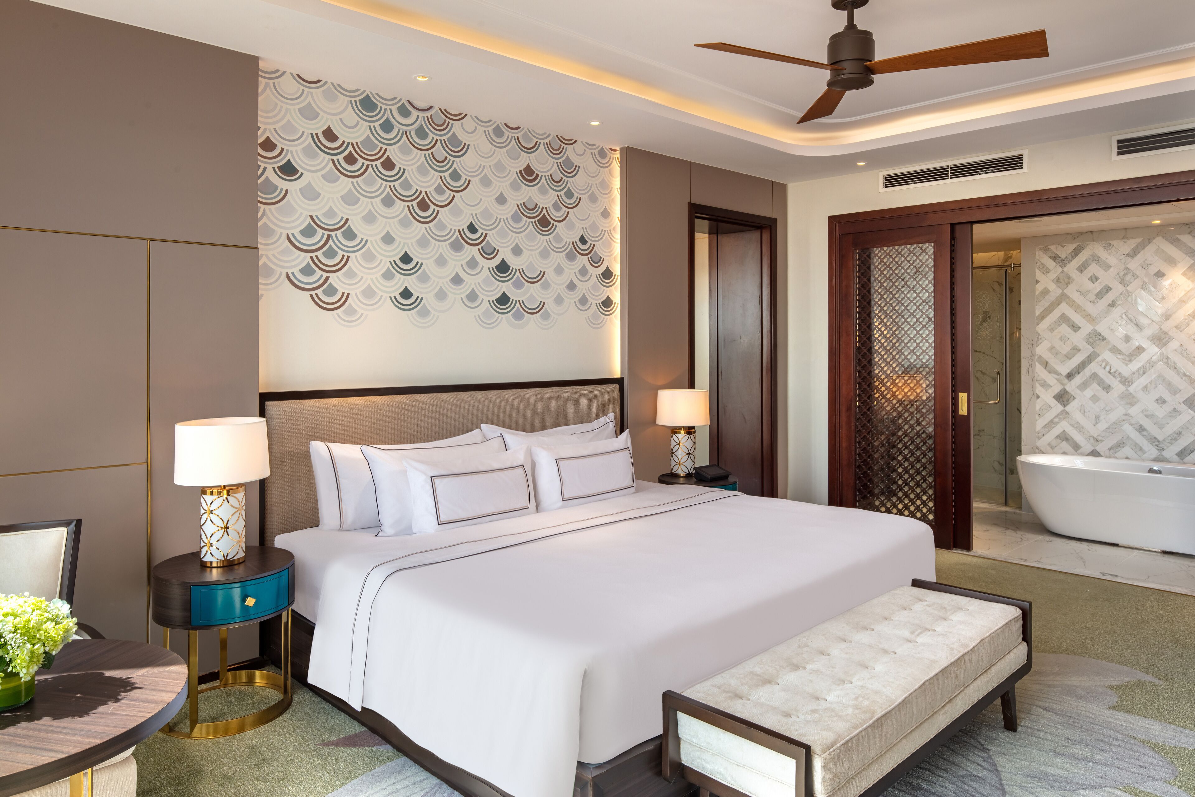 Une chambre d’hôtel moderne dotée d’un grand lit, de deux tables de chevet avec lampes, d’un banc, d’un ventilateur de plafond et d’une salle de bain avec baignoire.