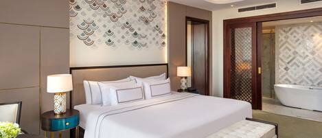 A modern hotel room with a large bed, two bedside tables with lamps, a bench, a ceiling fan, and a bathroom with a bathtub.