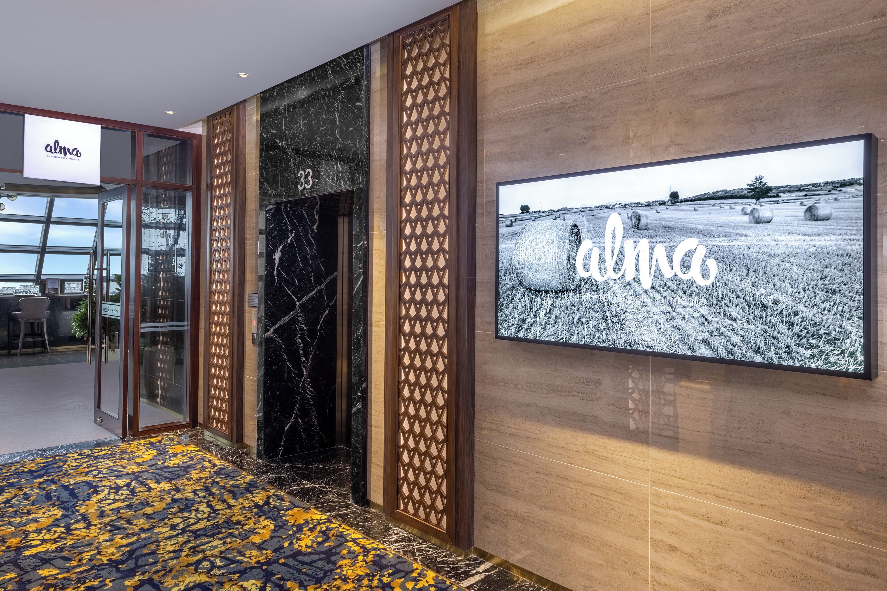 A hotel lobby with a modern design, featuring a large screen displaying the hotel's name, a patterned carpet, and a wooden decorative panel.