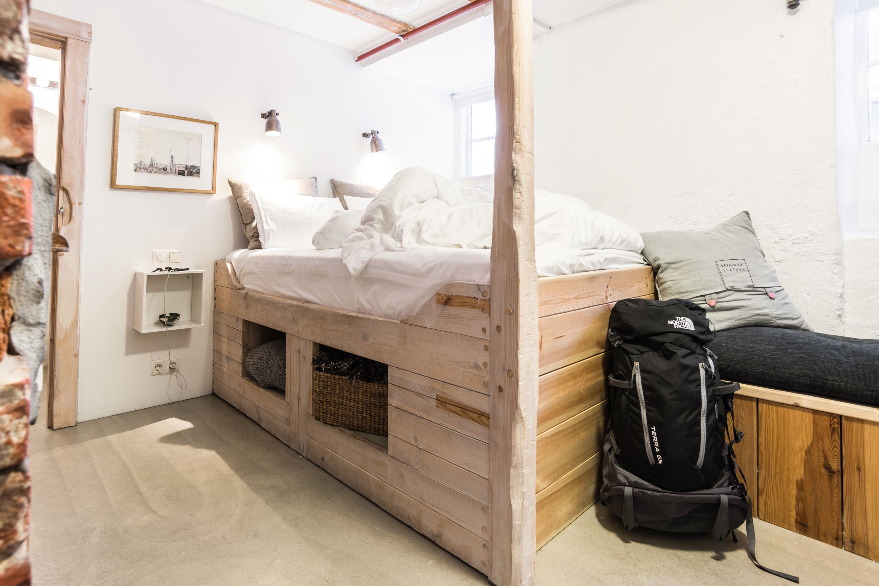 A room with a wooden bunk bed, a black backpack, a framed picture on the wall, and a small shelf with a basket.