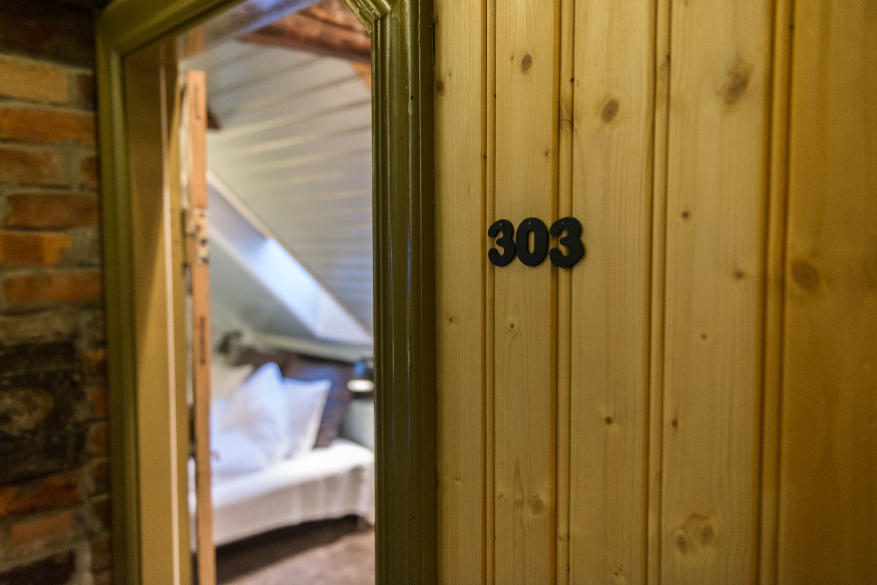 A wooden door with the number 303, a brick wall, and a glimpse of a bed in the background.