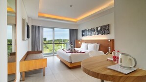A hotel room with a bed, a desk, a round table with a kettle, and a large window with a view of greenery. - Kuta Galleria Hotel (Kuta)