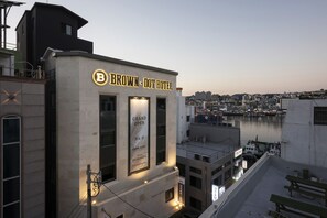 A hotel with a prominent 'B' logo and 'BROWN - DOT HOTEL' signage. It features a 'GRAND OPEN' banner and a view of the cityscape and water in the background. - Brown-Dot Tongyeong (Tongyeong)