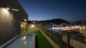 Una terraza en la azotea con piscina, mobiliario de exterior y vistas a un paisaje urbano al atardecer.