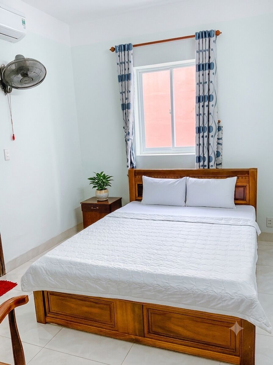 A bedroom with a wooden bed, a bedside table with a plant, a standing fan, and a window with curtains.