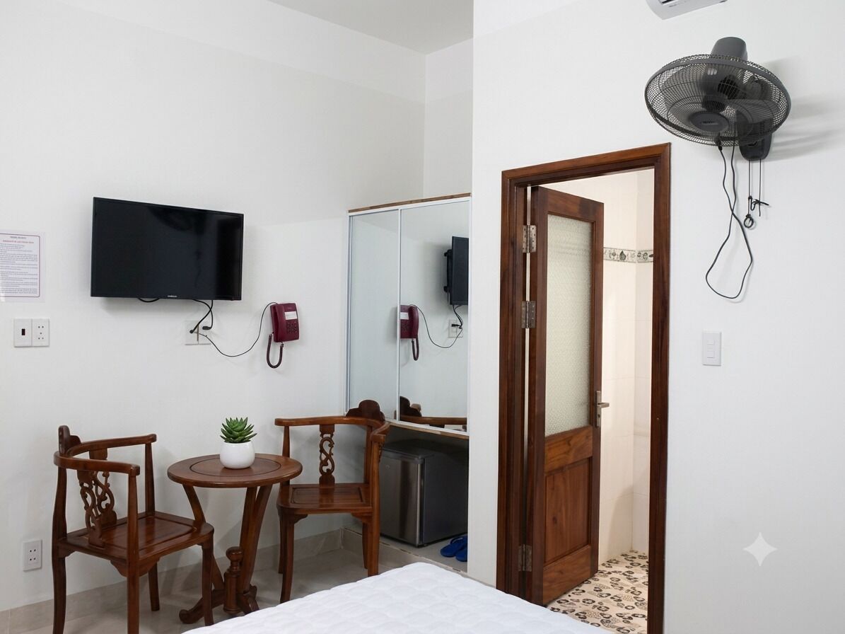 A hotel room with a bed, a TV mounted on the wall, a desk with a chair, a small table with a plant, a mirror, a refrigerator, and a wall-mounted fan.