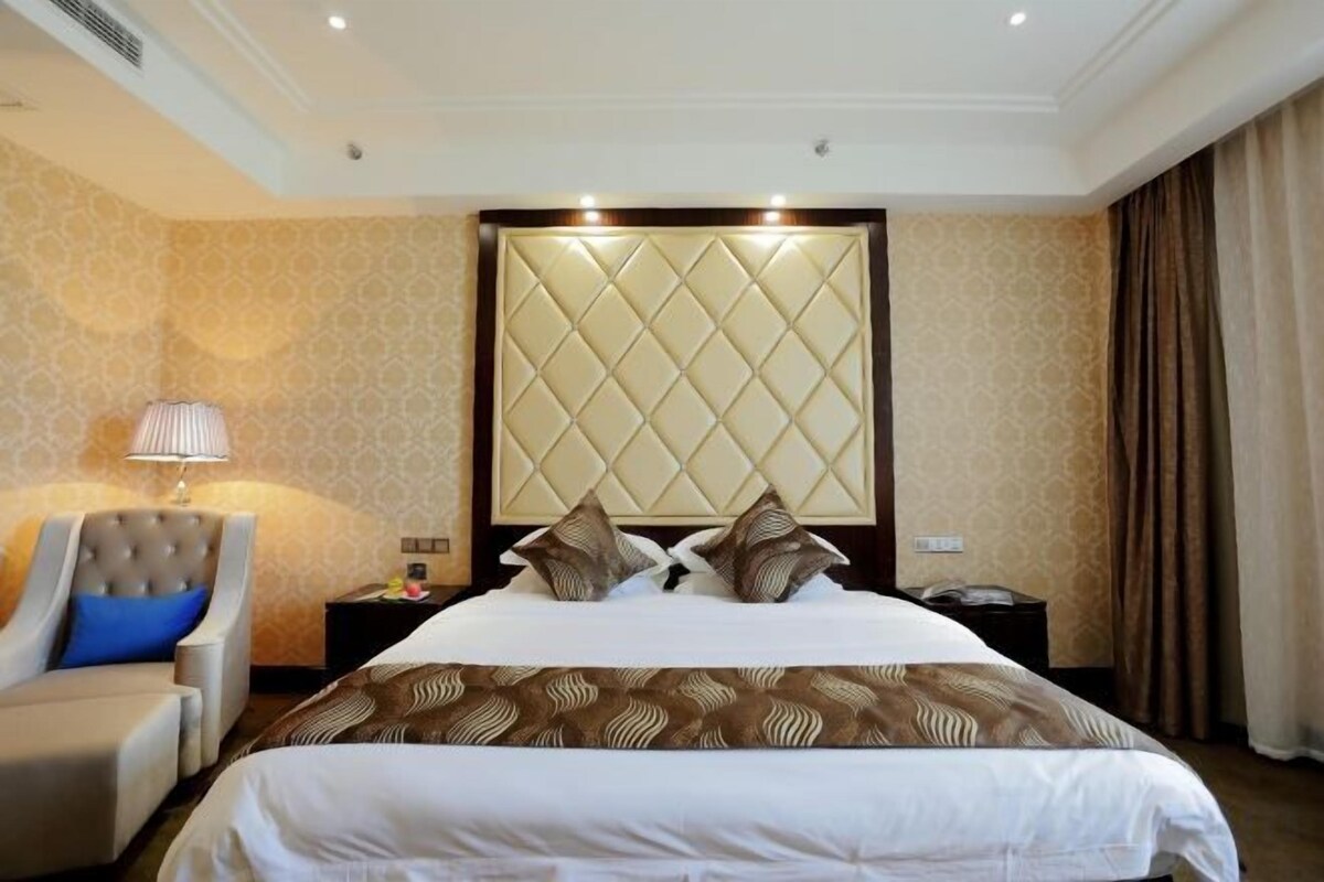 A hotel room with a large bed, a headboard with a quilted design, a bedside table with a lamp, and a chair.