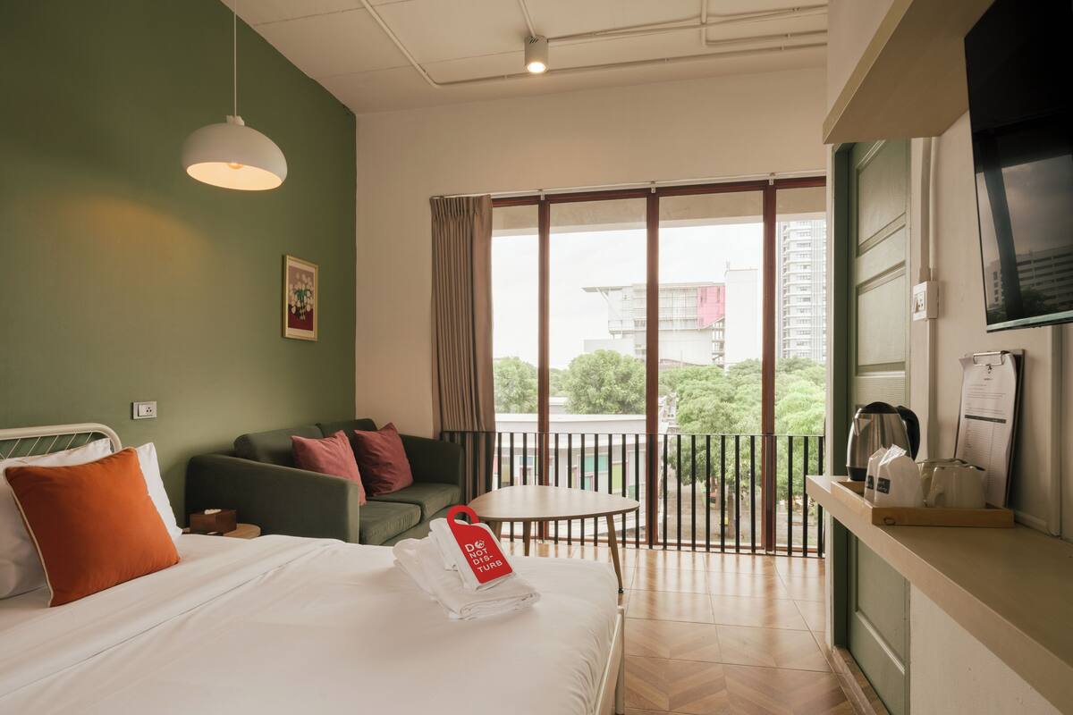 Hotelkamer met een bed, een bank, een tafeltje, een balkon met leuning en uitzicht op gebouwen en bomen.