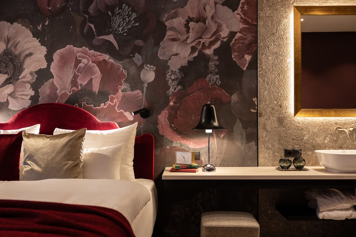 A hotel room with a bed, a bedside table with a lamp, a mirror, and a bathroom with a sink and towel rack.
