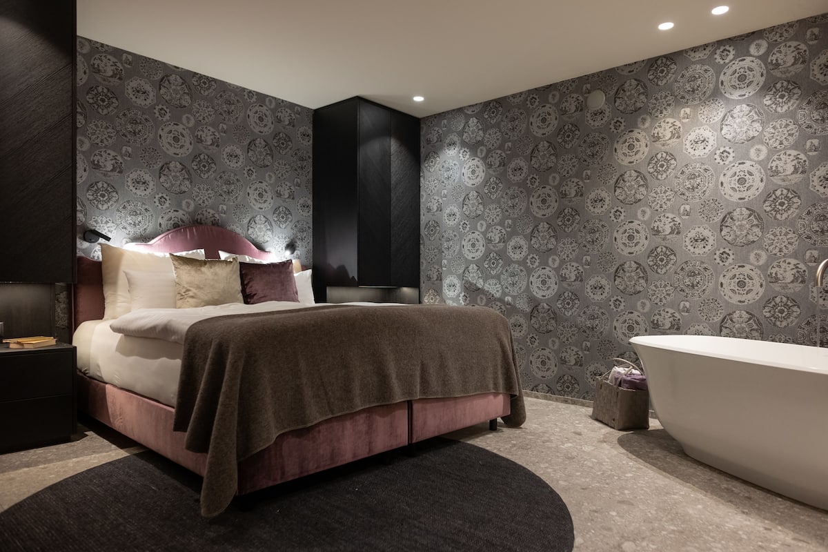 A modern bedroom with a large bed, a dark headboard, a vanity with a mirror, and a freestanding bathtub.