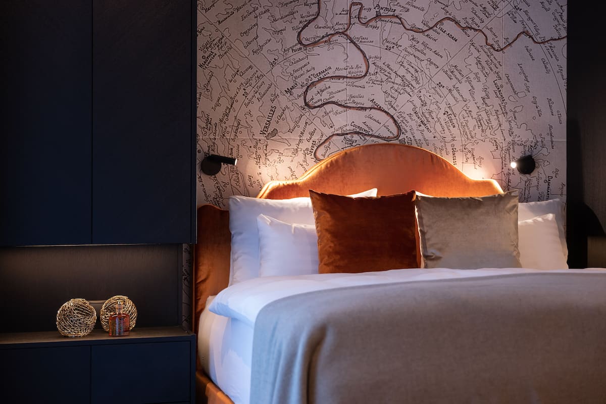 A neatly made bed with white linens and brown pillows, a dark wooden headboard, and a wall with a map design.