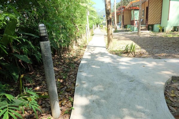 A concrete pathway flanked by dense foliage and a building with a tiled roof. - Khaosok Bamboo Huts Resort (Phanom)