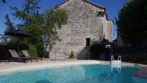 A swimming pool with lounge chairs and umbrellas, a stone building with a window, and a clear blue sky.