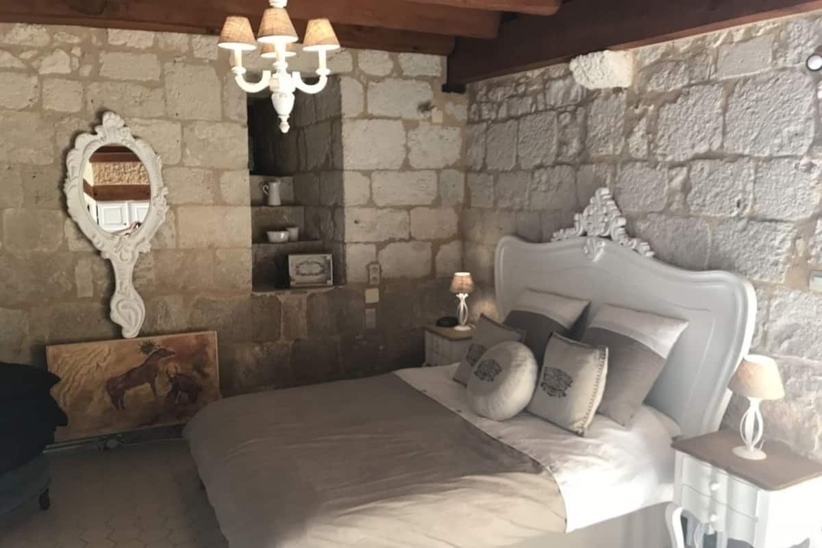 Ein Schlafzimmer mit Steinwand, einem weißen Bett mit grauer Bettwäsche, einem Holz-Kopfteil, einem Nachttisch mit Lampe und einem dekorativen Spiegel.