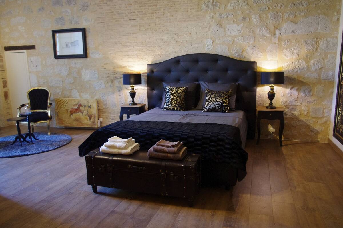 Ein Schlafzimmer mit einem großen Bett, zwei Nachttischen mit Lampen, einem Sessel, einem gerahmten Bild an der Wand und einem Koffer auf dem Boden.