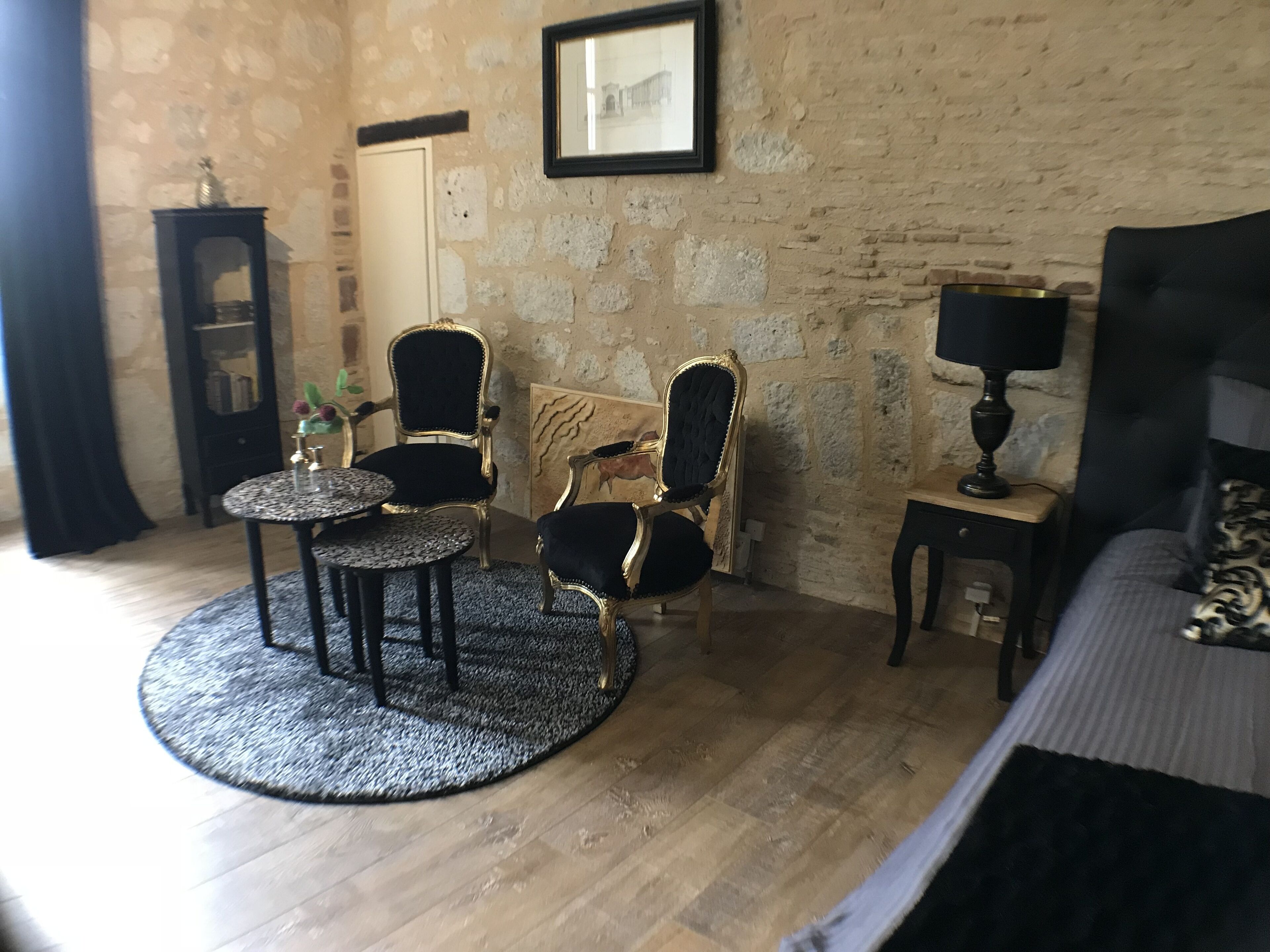 A room with a stone wall, a bed, a round table with chairs, a small cabinet, and a framed picture on the wall.