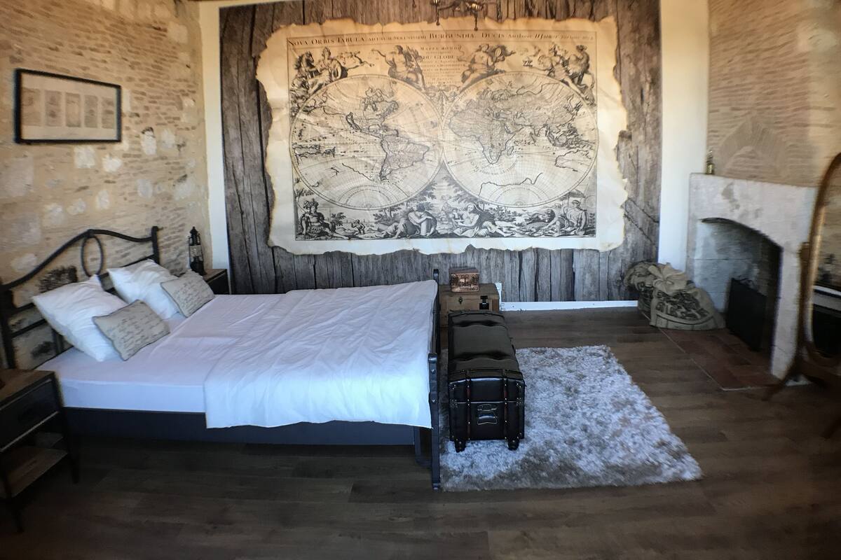 Ein Schlafzimmer mit einem großen Bett, einem hölzernen Kopfteil, einer Wandteppich-Karte, einem Kamin und einem Koffer auf einem Teppich.