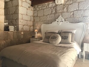 A bedroom with a stone wall, a white ornate headboard, and a bed with beige bedding and decorative pillows. - Château de Bouniagues (Bouniagues)