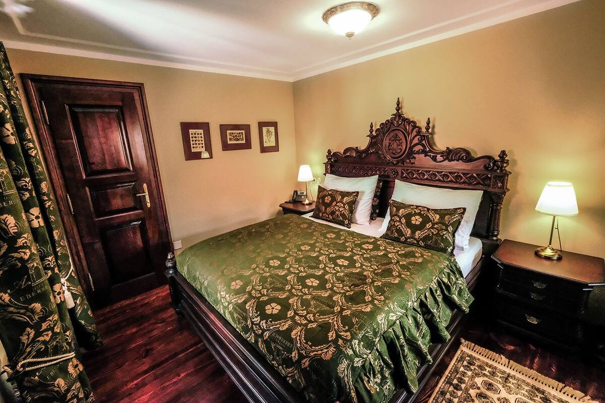 A bedroom with a large bed, ornate headboard, two bedside tables with lamps, a wooden door, and a patterned curtain.